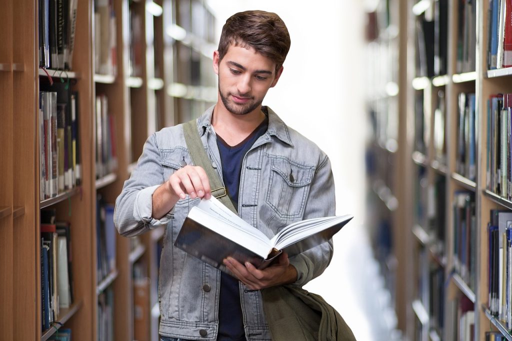 young man paging through a textbook by a library shelf